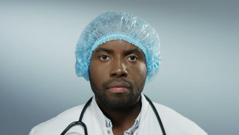 Close-up-of-the-young-African-American-handsome-man-doctor-looking-straight-to-the-camera-with-a-serious-face-on-the-grey-wall-background.-Portrait.